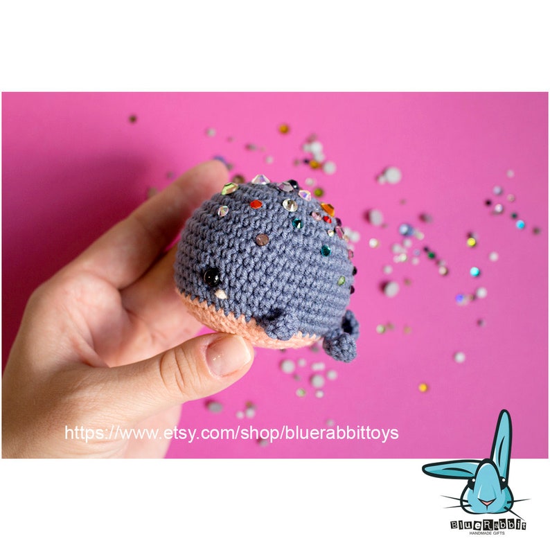 May include: A small crocheted whale toy with a pink belly and a blue body. The whale is covered in colorful rhinestones. The whale is being held in a hand against a pink background.