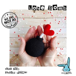 Puede incluir: Una esfera negra de ganchillo con forma de bomba, una mecha blanca y un coraz&oacute;n rojo de ganchillo en el extremo. El texto LOVE BOMB aparece arriba y el texto .PDF file crochet pattern abajo, con un sello de WE ARE NOT AI.