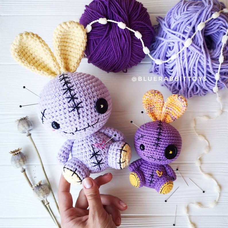 May include: Two purple crocheted bunny dolls with yellow ears and black button eyes. The dolls are stitched with black thread and have safety pins stuck in them. The dolls are sitting on a white surface with white yarn and purple yarn balls.