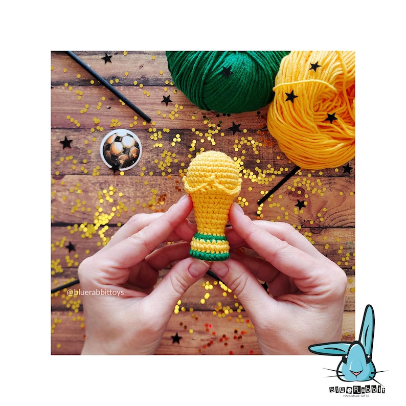 May include: A handmade crochet trophy in yellow, green, and gold is held by hands. The trophy is on a wooden surface with yarn, confetti, and a small soccer ball. The trophy is approximately 7.6 cm tall.