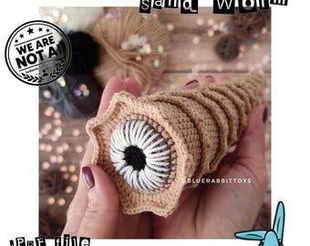 May include: A tan crochet sand worm toy with a star-shaped opening featuring a white and black centre. The text "sand worm" appears at the top, and "PDF file crochet pattern" is at the bottom. A "WE ARE NOT AI" logo is visible.