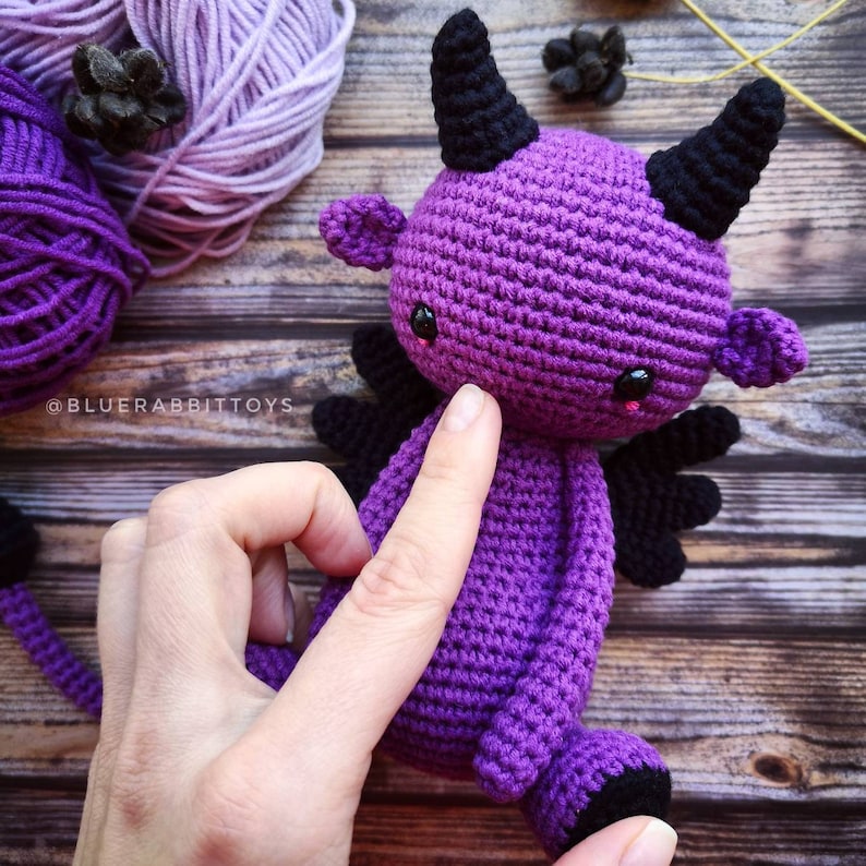 Puede incluir: Juguete de amigurumi de crochet morado con cuernos, ojos y alas negros. El juguete est&aacute; sentado sobre una superficie de madera.