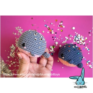 May include: Two gray crocheted whale plushies with pink bellies and sparkly rhinestones. The whales are sitting on a pink surface with scattered rhinestones.
