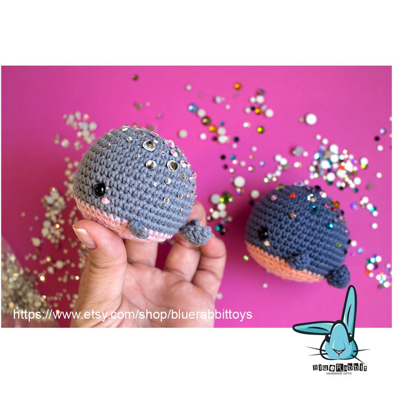 May include: Two gray crocheted whale plushies with pink bellies and sparkly rhinestones. The whales are sitting on a pink surface with scattered rhinestones.