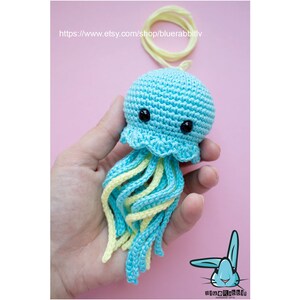 May include: A crocheted jellyfish toy featuring a rounded light blue head with black bead eyes and a scalloped edge. The jellyfish has multiple dangling tentacles in alternating light blue and pale yellow yarn, with a matching yellow hanging loop.