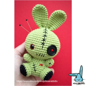 May include: Green crocheted bunny doll with black button eyes, a red button nose, and black stitching. The doll has black yarn for feet and is decorated with black stitching and safety pins.