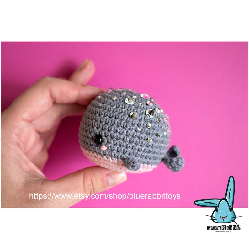 May include: A gray crocheted whale with pink accents and sparkly rhinestones. The whale has a small black eye and a small pink mouth. The whale is being held in a hand against a pink background.