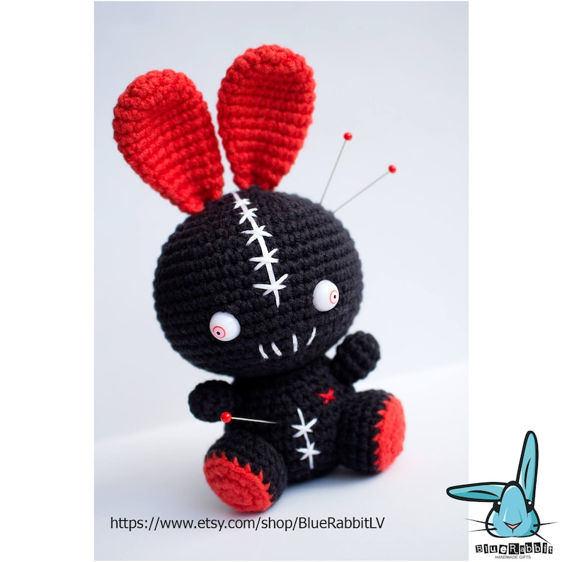 May include: Black crocheted bunny with red ears and white stitching. The bunny has googly eyes and is decorated with safety pins.