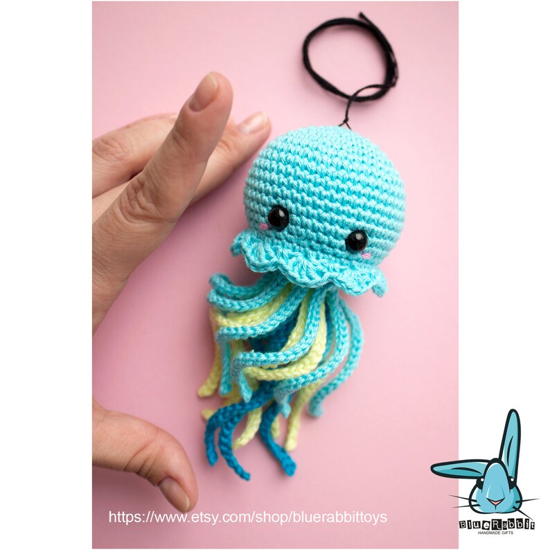 May include: A small, crocheted jellyfish ornament featuring a light blue rounded head with black safety eyes and pink stitched cheeks. It has a ruffled edge and dangling tentacles in alternating shades of light blue, yellow, and dark blue.