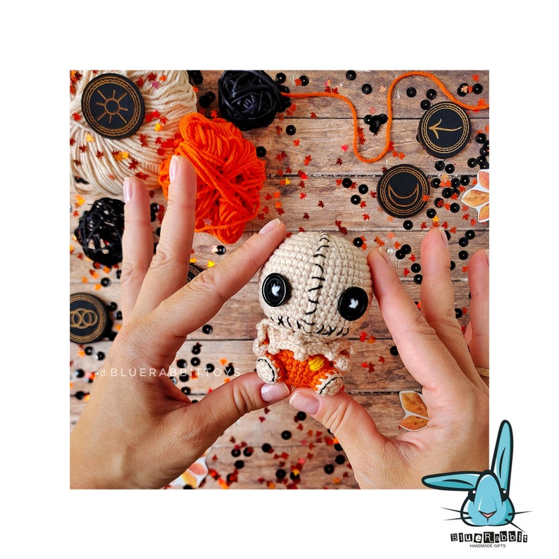 May include: A crocheted doll with a stitched-up mouth, button eyes, and orange pants. The doll is being held by two hands against a background of orange and black yarn, wooden beads, and fall leaves.
