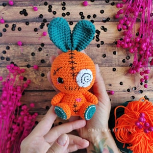 May include: A crocheted carrot-shaped toy with a stitched-on face, one eye is a white swirl, the other is black. The toy has teal bunny ears and is held in a person's hands.