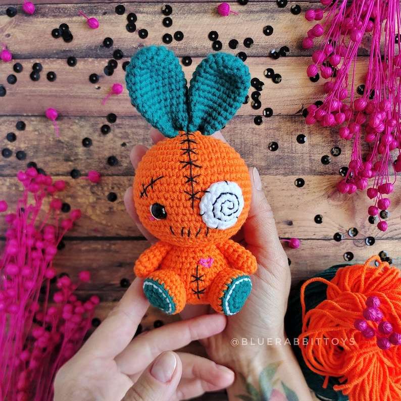 May include: A crocheted carrot-shaped toy with a stitched-on face, one eye is a white swirl, the other is black. The toy has teal bunny ears and is held in a person's hands.