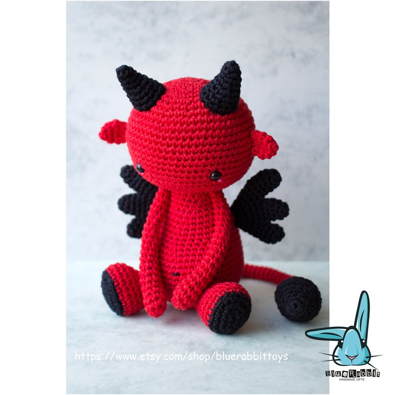 Puede incluir: Un juguete de peluche de diablo rojo tejido a crochet con cuernos, alas y cola negros. El juguete tiene una cara linda y de dibujos animados con ojos negros y una peque&ntilde;a boca.