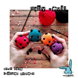 以下が含まれることがあります： 赤、ティール、オレンジ、紫の4つの編みぐるみの悪魔のおもちゃ。それぞれ黒い角と笑顔の顔があります。「Baby Devil」の文字がおもちゃの上に、「PDF file crochet pattern」が下にあります。おもちゃは手で持たれています。