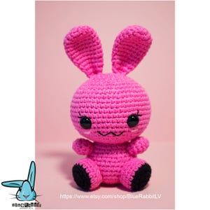 May include: A pink crocheted bunny with black eyes and a black nose. The bunny is sitting on a pink surface.