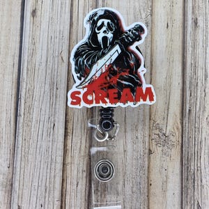 May include: A retractable badge reel featuring a graphic of the Scream mask holding a knife with the word "SCREAM" in red. The reel has a clear plastic strap and a metal clip.