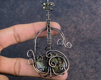 May include: A copper wire pendant, crafted to resemble a musical instrument. It features a central oval stone with green and brown tones. The design incorporates decorative swirls and small brass-coloured beads.