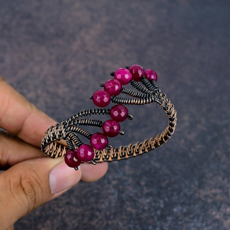 May include: A wire-wrapped cuff bracelet featuring dark oxidised copper wire and multiple faceted, round, deep magenta-coloured beads. The design incorporates a spiralling structure with beads attached to individual wire stems along the top edge.