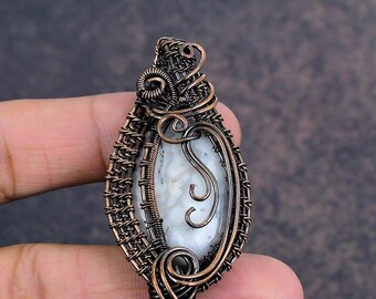 Peut inclure: Un pendentif artisanal avec un centre en pierre blanche, enfermé dans un fil de cuivre complexe. Le filigrane présente des motifs tissés et enroulés, créant un design artistique unique. La pierre a un aspect marbré.
