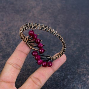 May include: A wire-wrapped cuff bracelet crafted from dark oxidised copper wire, featuring a series of small, round, deep magenta-coloured beads arranged in a curved, branch-like pattern along the inner edge of the circular metal frame.