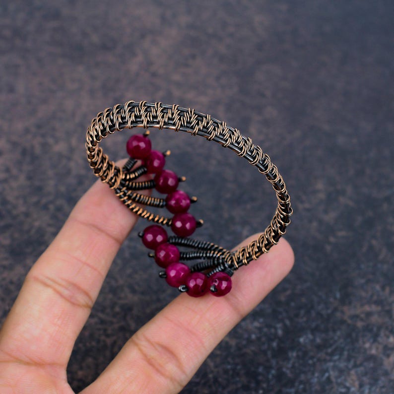 May include: A wire-wrapped cuff bracelet crafted from dark oxidised copper wire, featuring a series of small, round, deep magenta-coloured beads arranged in a curved, branch-like pattern along the inner edge of the circular metal frame.