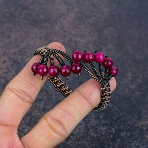 May include: A wire-wrapped cuff bracelet crafted from dark oxidised copper wire, featuring a series of faceted, round, deep magenta-coloured beads arranged in clusters along the top edge of the band, creating a textured and layered appearance.