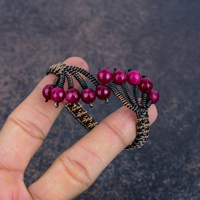 May include: A wire-wrapped cuff bracelet crafted from dark oxidised copper wire, featuring a series of faceted, round, deep magenta-coloured beads arranged in clusters along the top edge of the band, creating a textured and layered appearance.