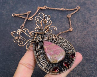 Puede incluir: Collar con colgante envuelto en alambre de cobre con una gema rosa grande y tres gemas rojas más pequeñas. El colgante tiene un intrincado diseño tejido y una cadena decorativa de cobre.