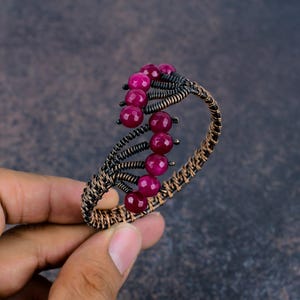 May include: A wire-wrapped cuff bracelet featuring dark oxidised copper wire and a series of faceted, round, deep magenta-coloured beads. The design incorporates intricate coiled wire patterns that secure the beads along the open-ended band.
