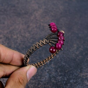 May include: A cuff bracelet crafted from dark oxidised copper wire woven into a structured band. The design features a series of faceted, deep magenta-coloured round beads arranged in a fan-like pattern along one side of the open-ended wire frame.