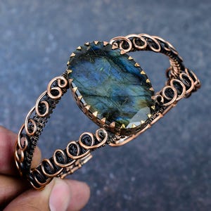 May include: A cuff bracelet featuring a large, oval-shaped labradorite gemstone with blue and green iridescence. The stone is set in a copper-toned metal frame, surrounded by intricate wire-wrapped detailing in dark and copper-colored metal.