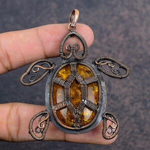 May include: A handcrafted turtle pendant with a copper-colored wire frame and a central amber-colored stone. The pendant features intricate wire detailing forming the turtle's shell, head, and flippers. The pendant is approximately 2 inches long and 1.5 inches wide.