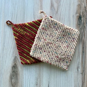 May include: Two handmade, square potholders. One is a mix of red, yellow, and green, the other is off-white with flecks of color. Both have a loop for hanging. The potholders are made of a woven material.