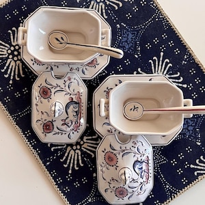 May include: Two cream-colored ceramic sugar bowls with matching lids and spoons, featuring hand-painted floral motifs and a small animal illustration in blue, pink, and brown tones. Each bowl has two side handles and sits on a matching rectangular saucer.