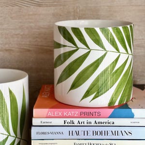 May include: Two white ceramic planters with green palm leaf designs. The planters are stacked on top of a stack of books. The top book has the text "ALEX KATZ PRINTS" and the text "Folk Art in America".