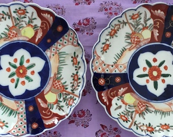 May include: Two decorative ceramic plates with scalloped edges. The plates feature a blue and white floral design with red, gold, and green accents. The plates are on a purple fabric background.
