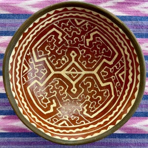 May include: A handmade ceramic bowl with a reddish-brown interior and a cream-colored geometric design. The rim is dark brown, and the bowl sits on a striped purple and blue fabric. This decorative bowl is perfect for serving food or as a decorative piece.