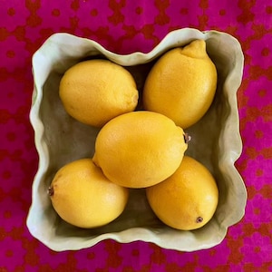 May include: A ceramic bowl with a ruffled edge, filled with five yellow lemons. The bowl is light green and has a matte finish. The lemons are arranged in a cluster, with some overlapping.
