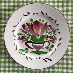 May include: A white ceramic plate with a hand-painted floral design. The central motif features a large, deep purple tulip with yellow accents, surrounded by green leaves and smaller flowers. A brown decorative border and a maroon rim complete the design.