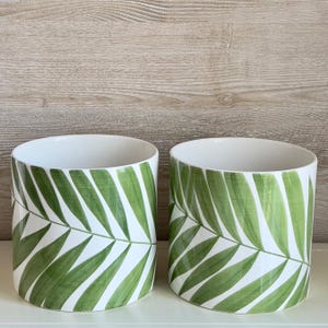 May include: Two white ceramic planters with a green palm leaf design. The planters are cylindrical and have a white background with green palm leaves.