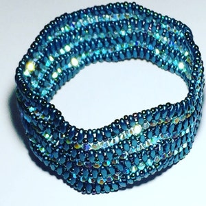 May include: A teal blue beaded bracelet with iridescent accents. The bracelet is made with small, round beads and has a woven design.