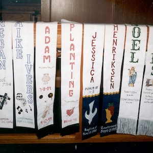 May include: White fabric banners with embroidered names, dates, and bible verses. The banners are hanging on a wooden railing.