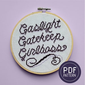 Peut inclure: Un cerceau de broderie blanc avec un motif de point de croix noir qui dit "Gaslight Gatekeep Girlboss". Le cerceau est en bois et a une finition naturelle. L'arrière-plan est violet clair.