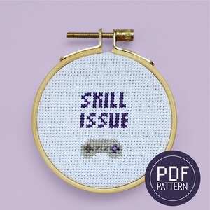 May include: A white cross-stitch embroidery hoop with a purple and gray design. The design includes the text "Skill Issue" and a stylized image of a video game controller. The hoop is labeled "PDF Pattern".