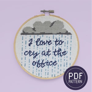 May include: A cross-stitch embroidery hoop with a blue and white design. The design features a gray cloud with raindrops and the text "I love to cry at the office".