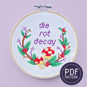 May include: A cross-stitch embroidery hoop with a white background and a purple text that reads "die rot decay" surrounded by a green wreath with red berries and mushrooms. The hoop is made of wood and has a metal ring.