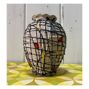 May include: A decorative ceramic vase with a cream base, overlaid with a black web-like pattern. The vase features colourful accents of red, yellow, and blue. The rim is a light green colour. The vase is on a yellow and green patterned surface.