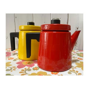 May include: Two vintage enamel coffee pots, one red and one yellow, with black handles. The pots are sitting on a floral patterned surface.