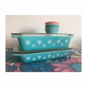 May include: A set of three turquoise blue casserole dishes with white snowflake designs. The dishes are stacked on top of each other. There are three matching turquoise blue bowls stacked on top of each other in the background.