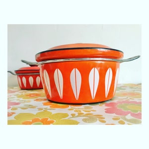 May include: Two orange enamel pots with white leaf-shaped patterns. The pots have silver handles and lids. The background features a floral design. These are retro kitchenware.